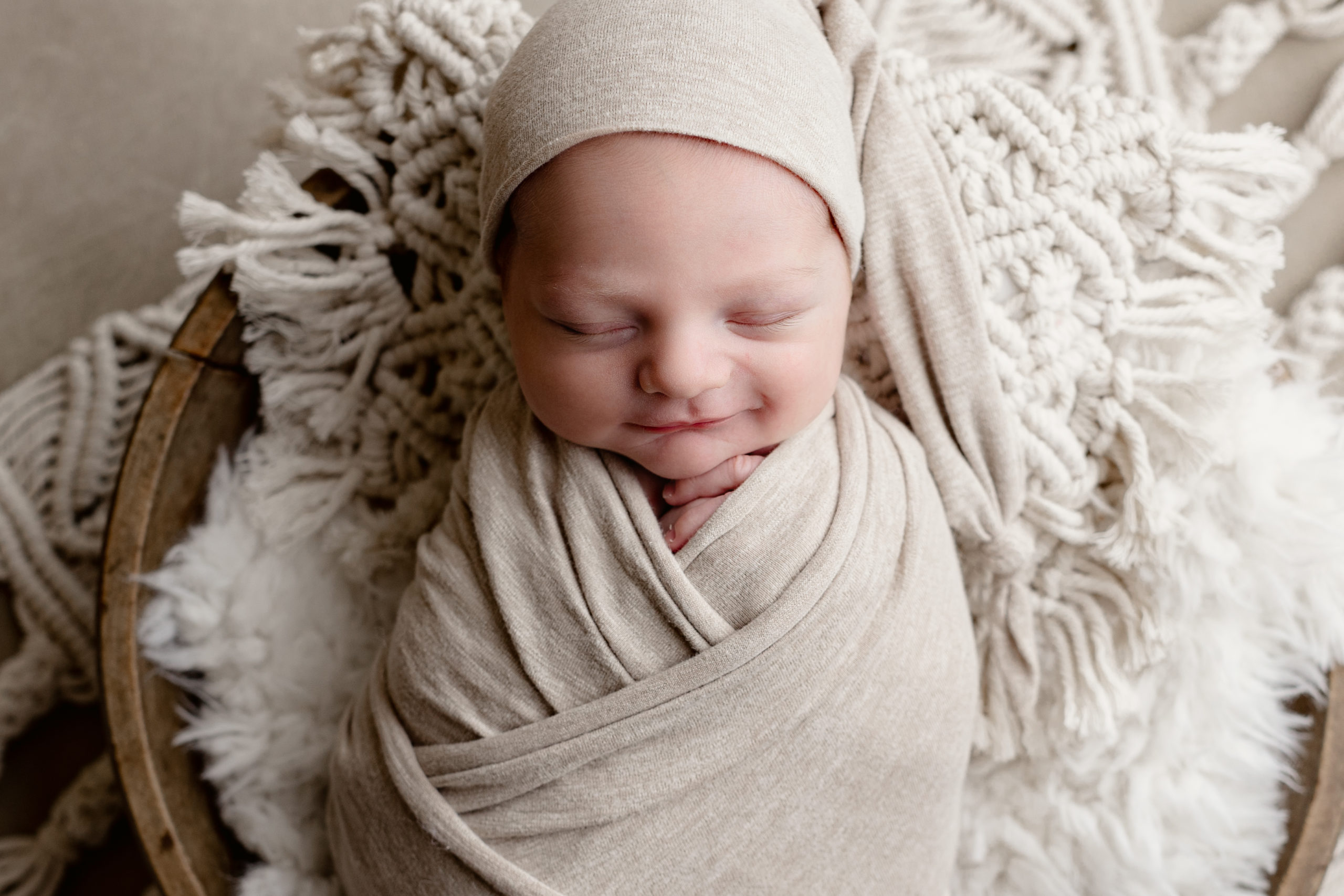 newborn baby boy smiling while wearing a tan sleepy cap with a tan blanket and macrame pillow during pictures with Milwaukee newborn photographer