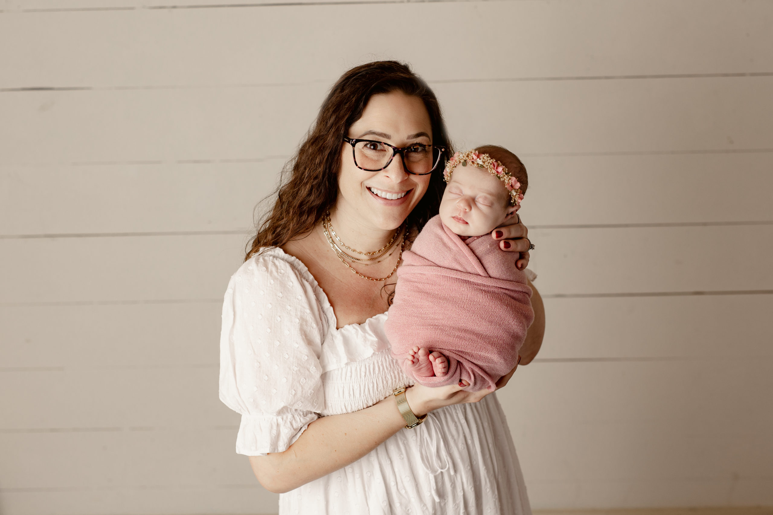 mom in white dress and glasses holding up her newborn baby girl in pink Mommy And Me Classes Milwaukee