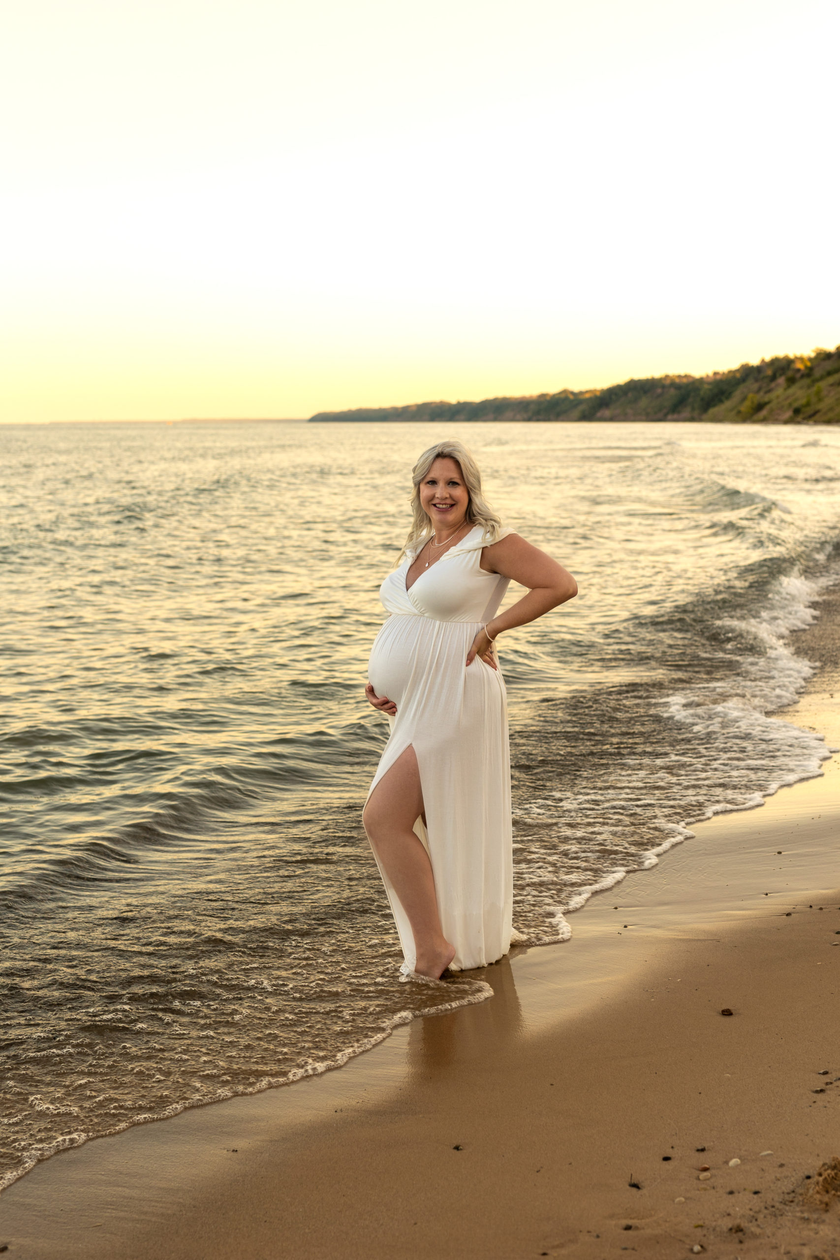 mom to be in white maternity gown standing near the edge of the water Vios Fertility Milwaukee