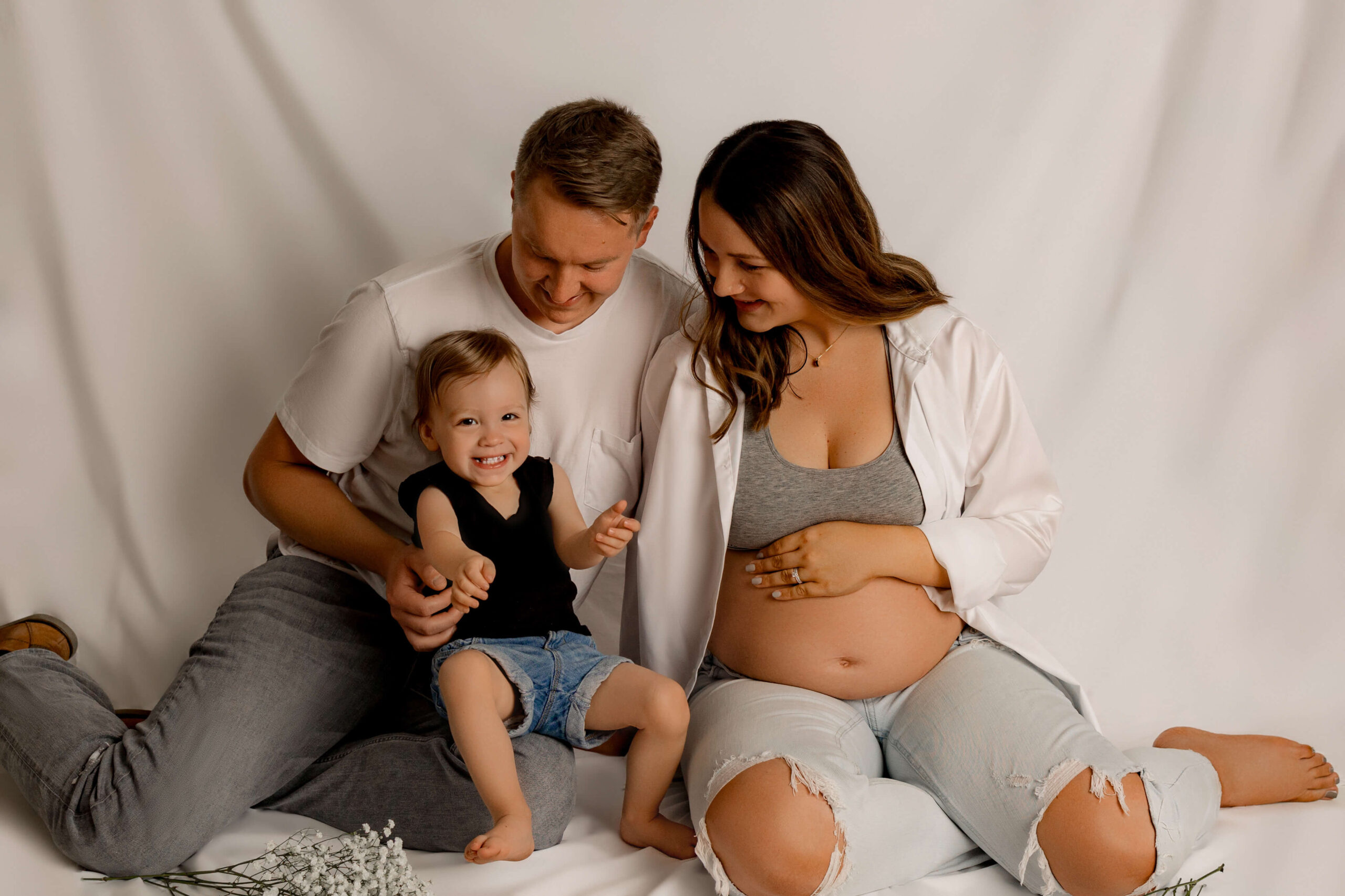 maternity photos with ripped jeans and belly showing on a white sheet backdrop