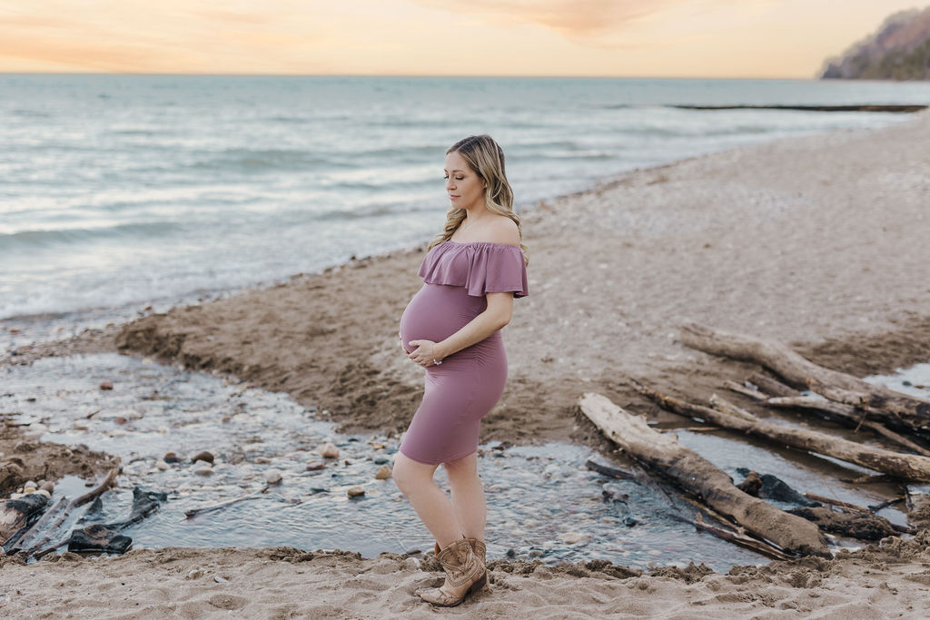 A mom to be in a pink maternity gown and cowboy boots stands on a beach at sunset while holding the bump