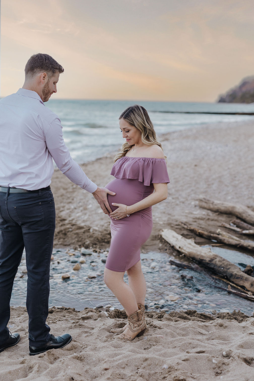 A mother to be stands on a beach in a pink maternity dress and cowboy boots while her husband holds the bump after getting 3d ultrasound milwaukee