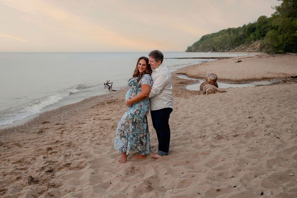A happy dad to be hugs his pregnant wife from behind as they stand on a beach at sunset