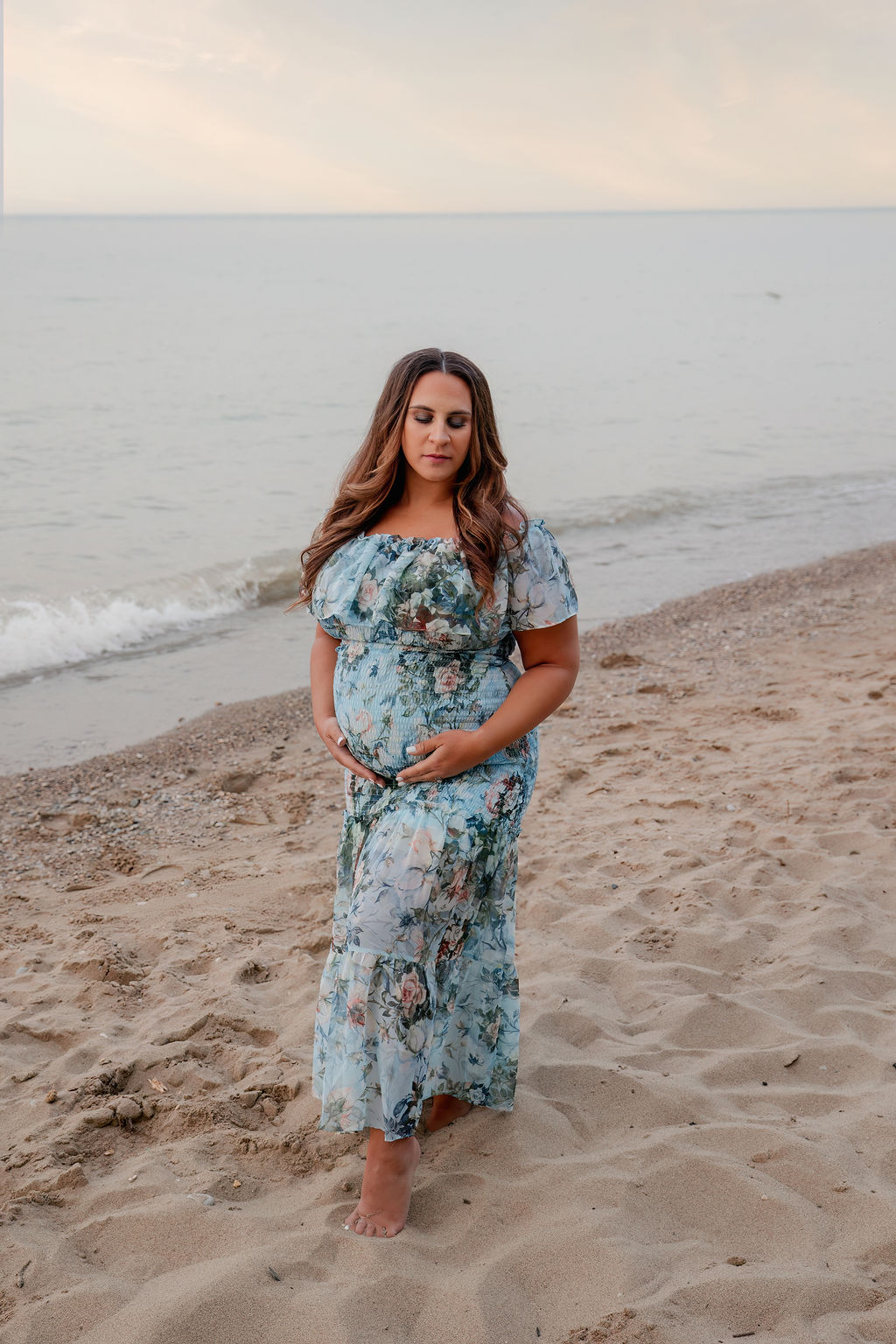 A mother to be in a blue floral print maternity dress walks down a beach at sunset holding her bump after visiting Froedtert birth center