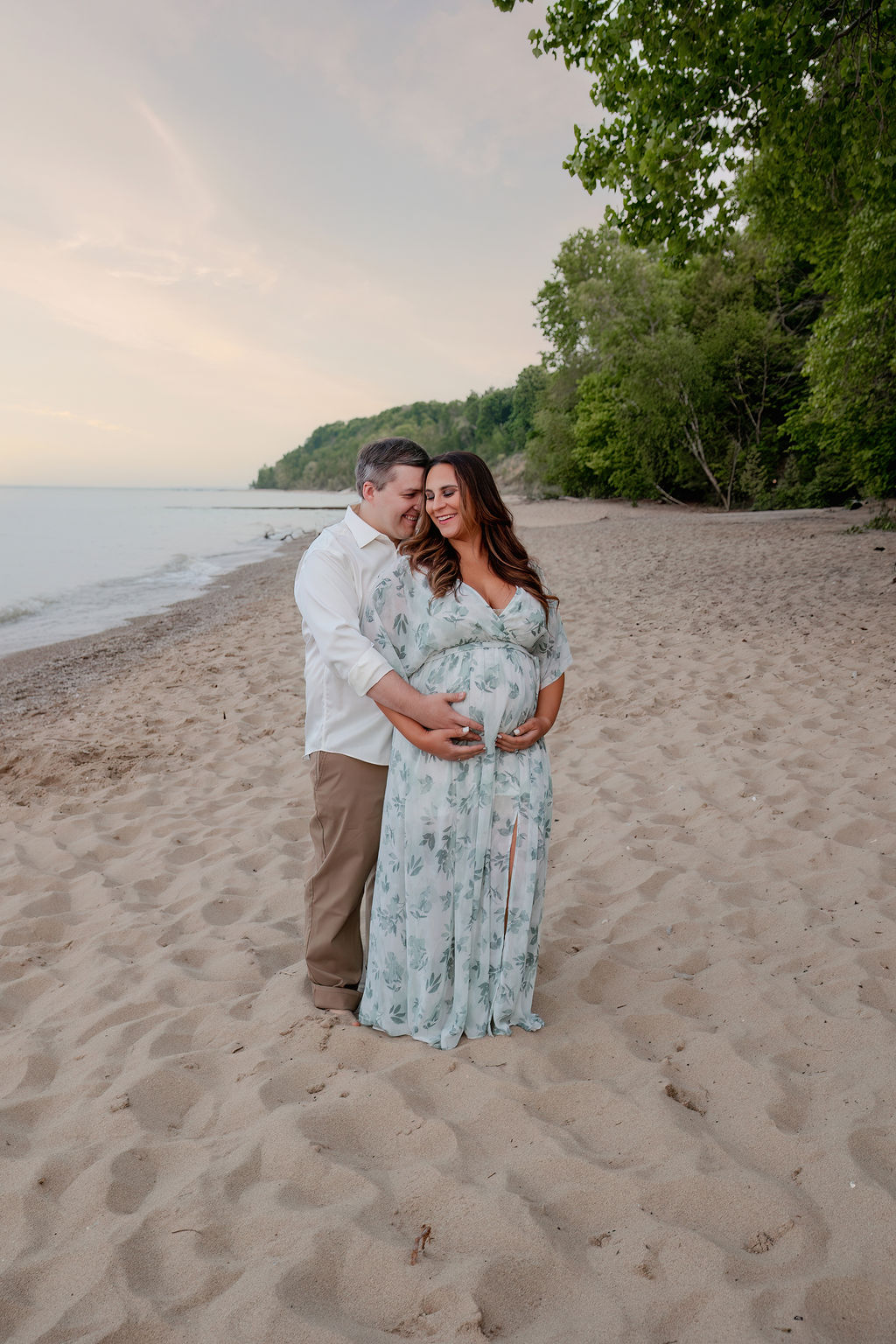 Happy expecting parents snuggle while standing on a beach in a blue floral maternity dress after visiting Froedtert birth center