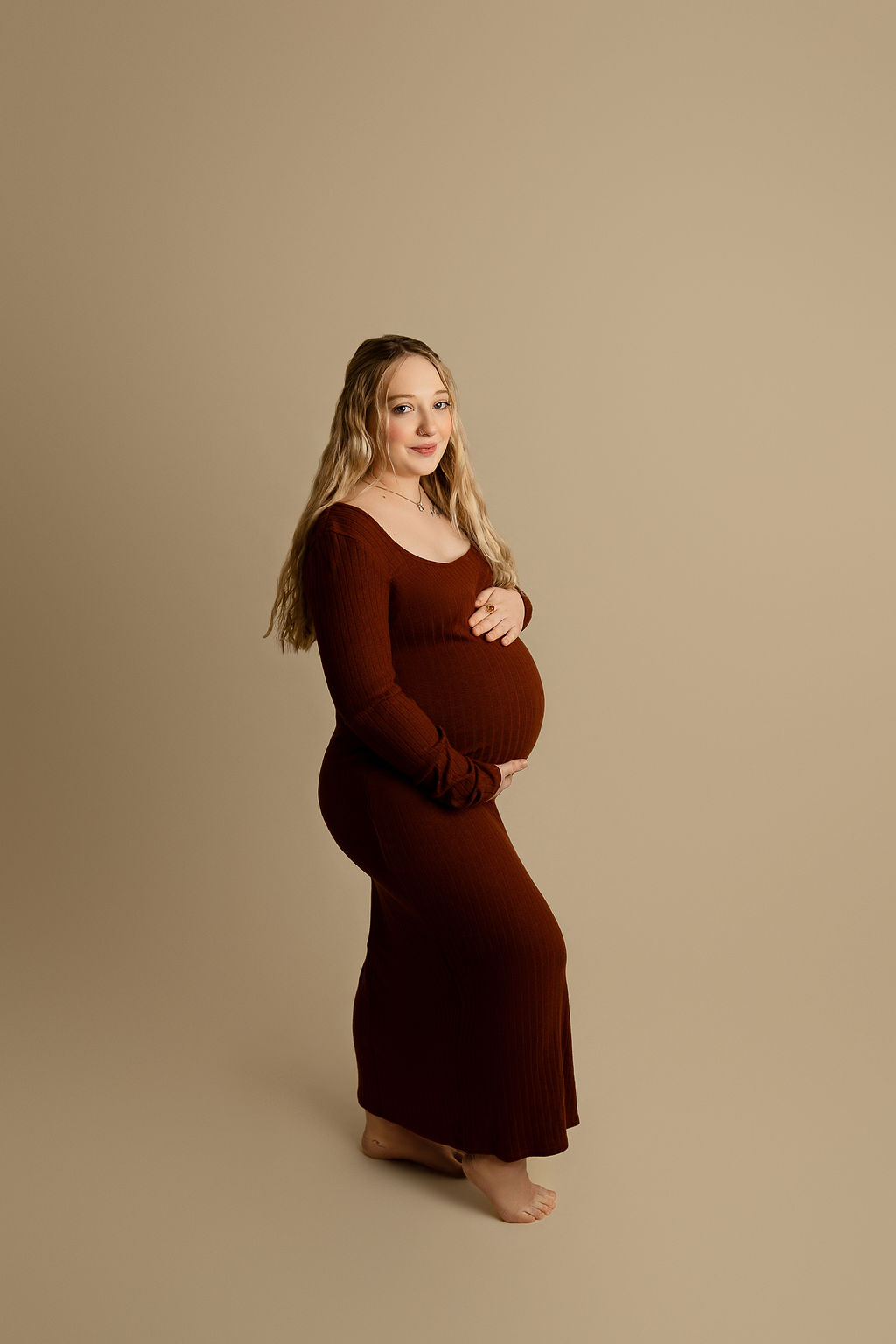 A blonde mother to be smiles while holding her bump and standing in a studio