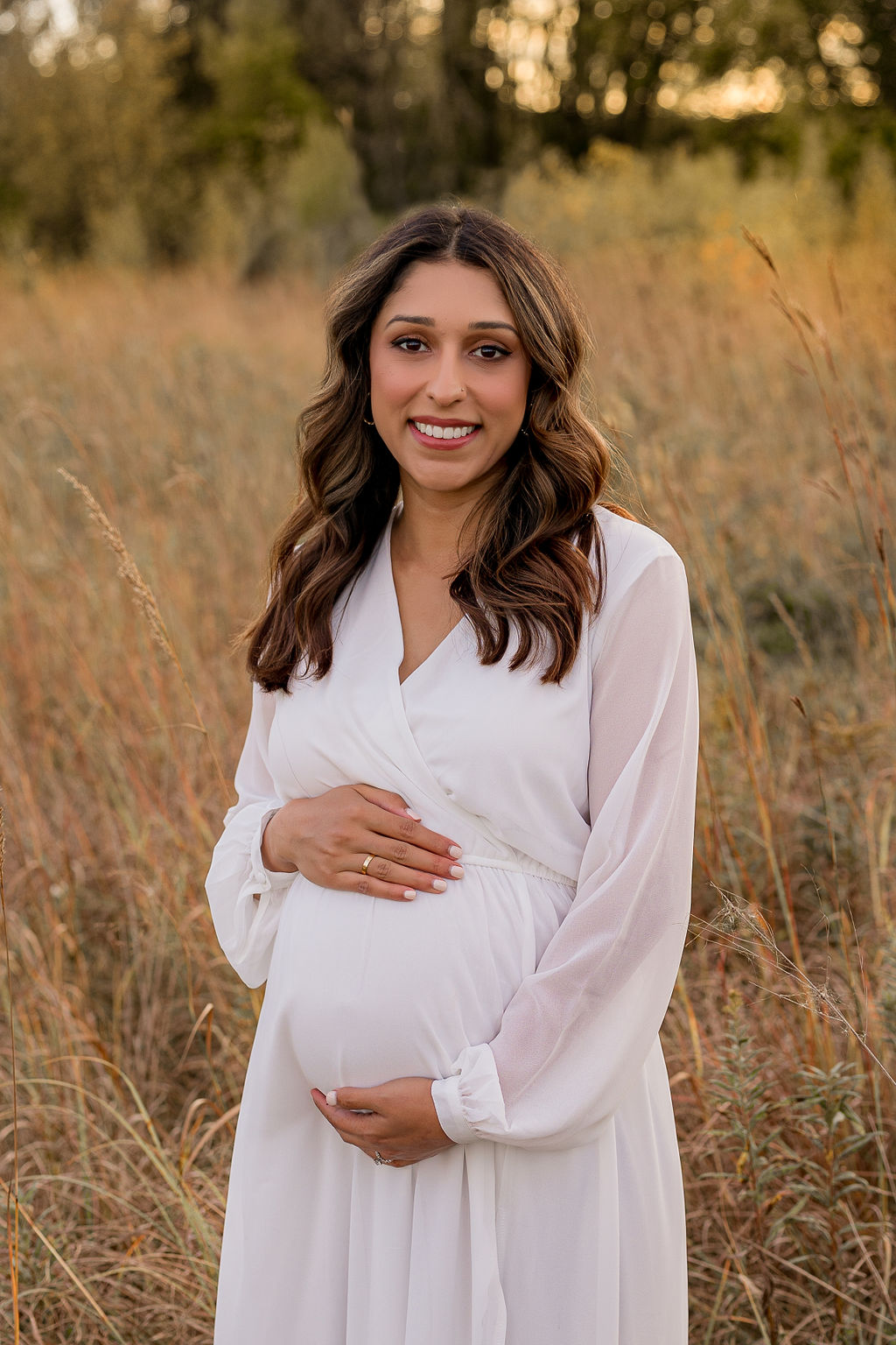 A happy mother to be in a white maternity gown holds her bump while standing in a golden field at sunset