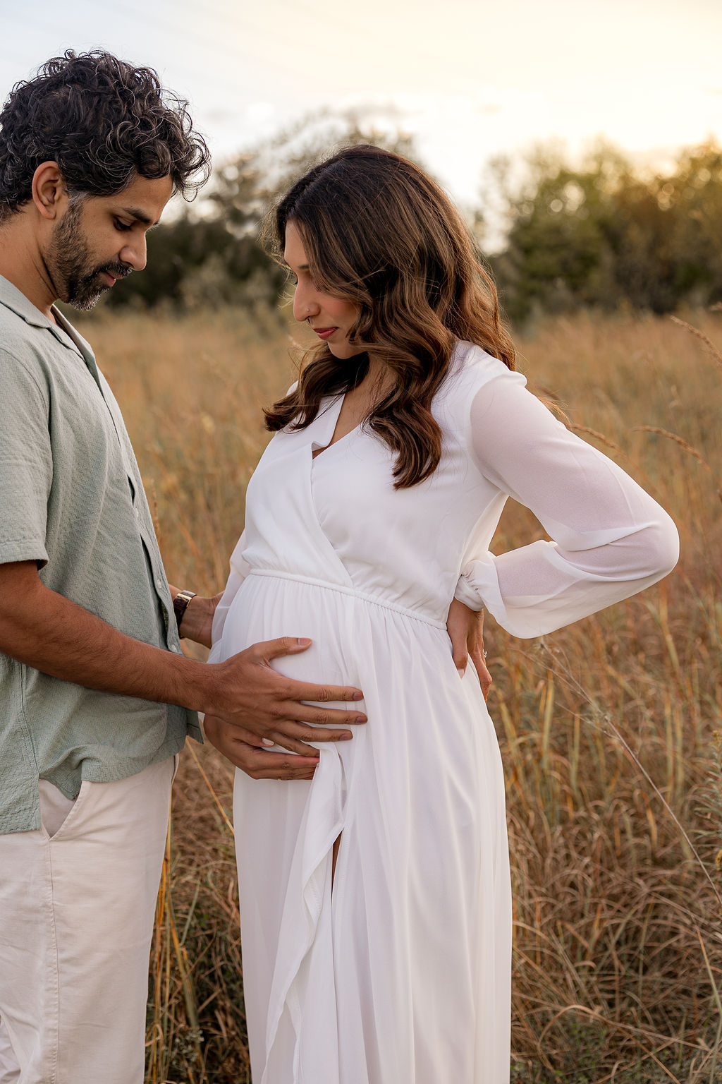 A mother and father to be stand in park field at sunset holding the bump in a white maternity dress after visiting authentic birth center