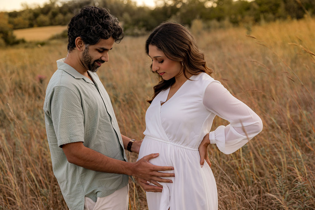 A happy father to be smiles while holding the bump in a field of tall golden grasses at sunset after visiting authentic birth center