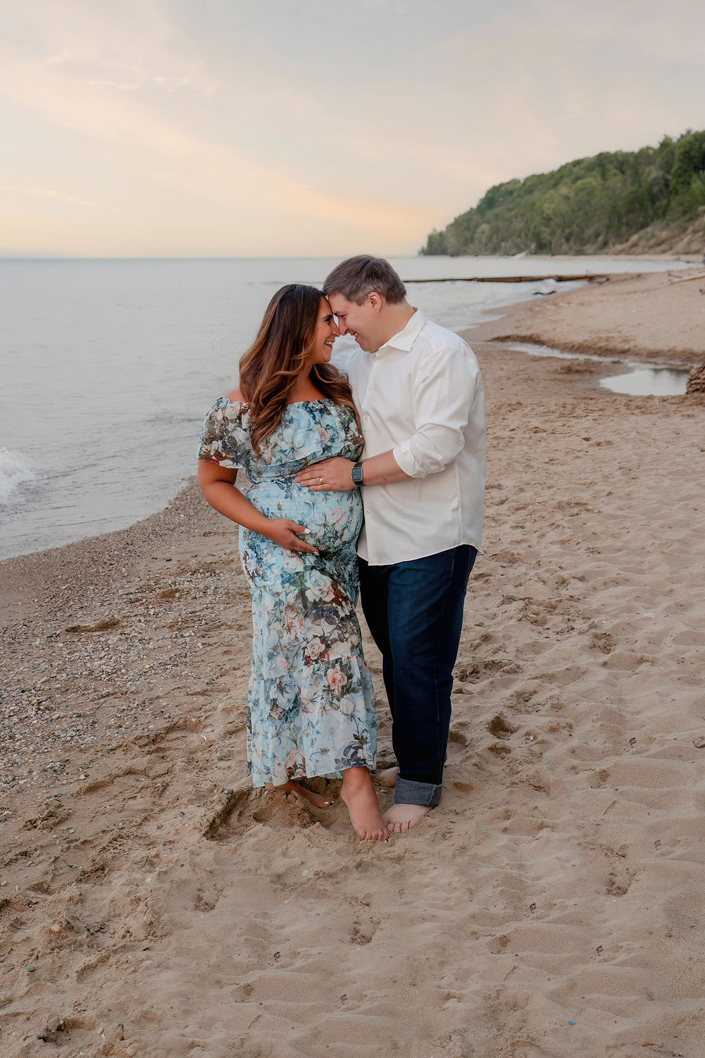 Expecting parents smile and touch foreheads and the bump while standing on a beach at sunset