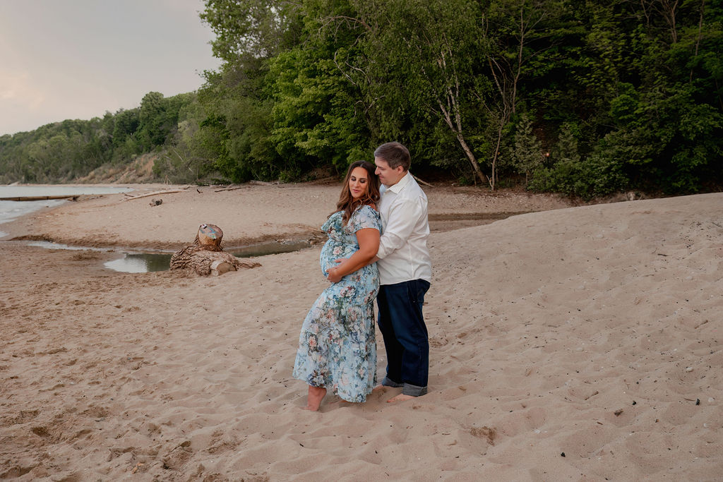 A mother to be leans into her husband as they both hold the bump in a blue floral print maternity gown and standing on a beach after visiting birthing centers in milwaukee wi