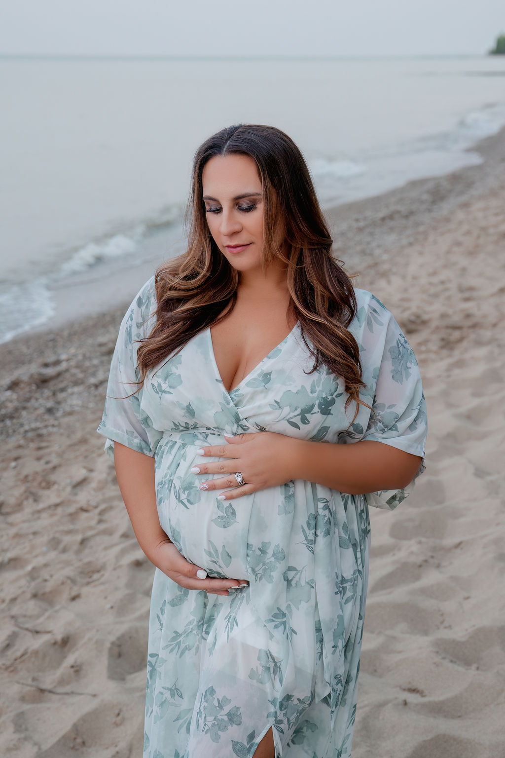 A mother to be in a blue floral print dress stands on a beach smiling down at her bump after visiting birthing centers in milwaukee wi
