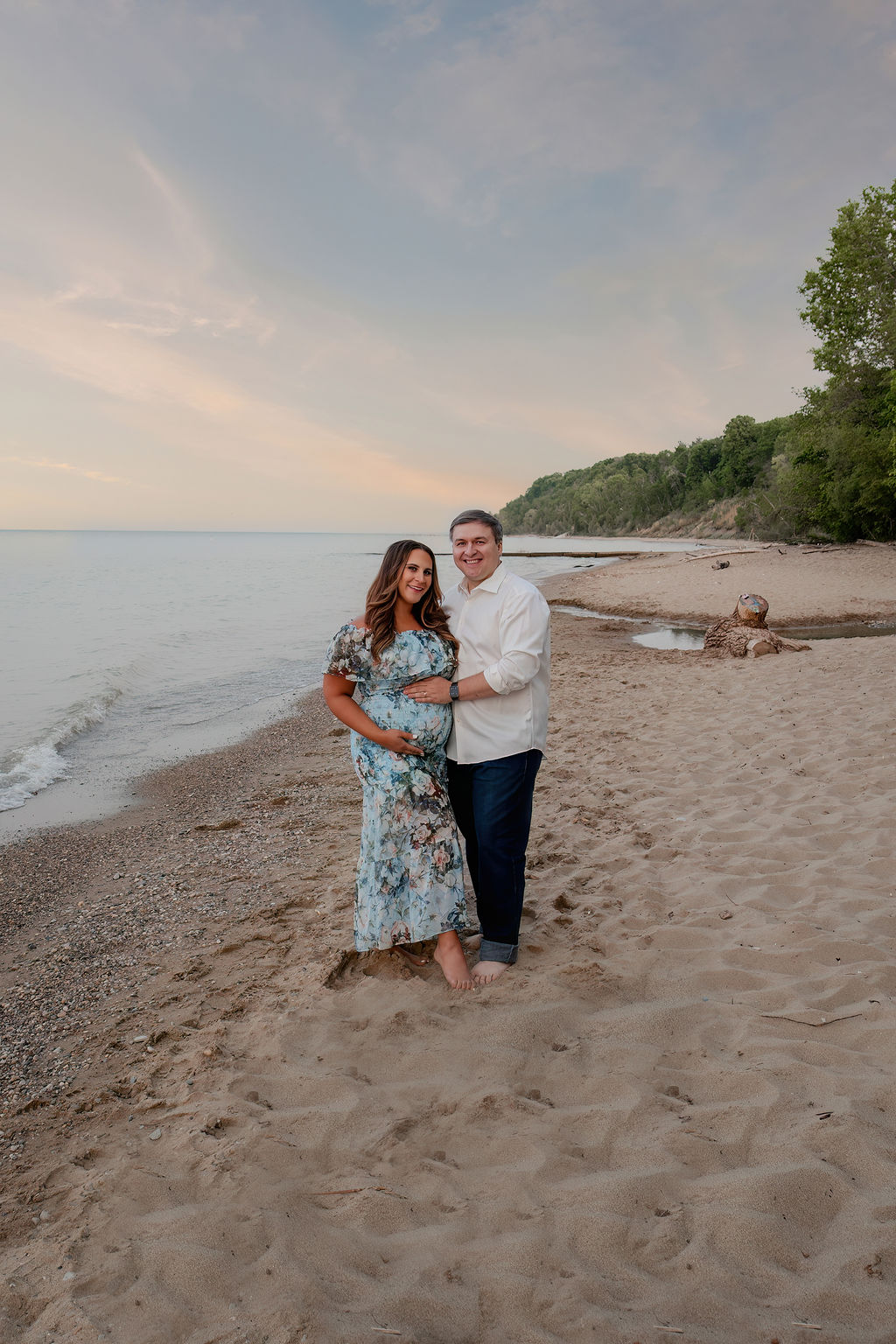 Happy expecting parents stand on a beach at sunset holding the bump