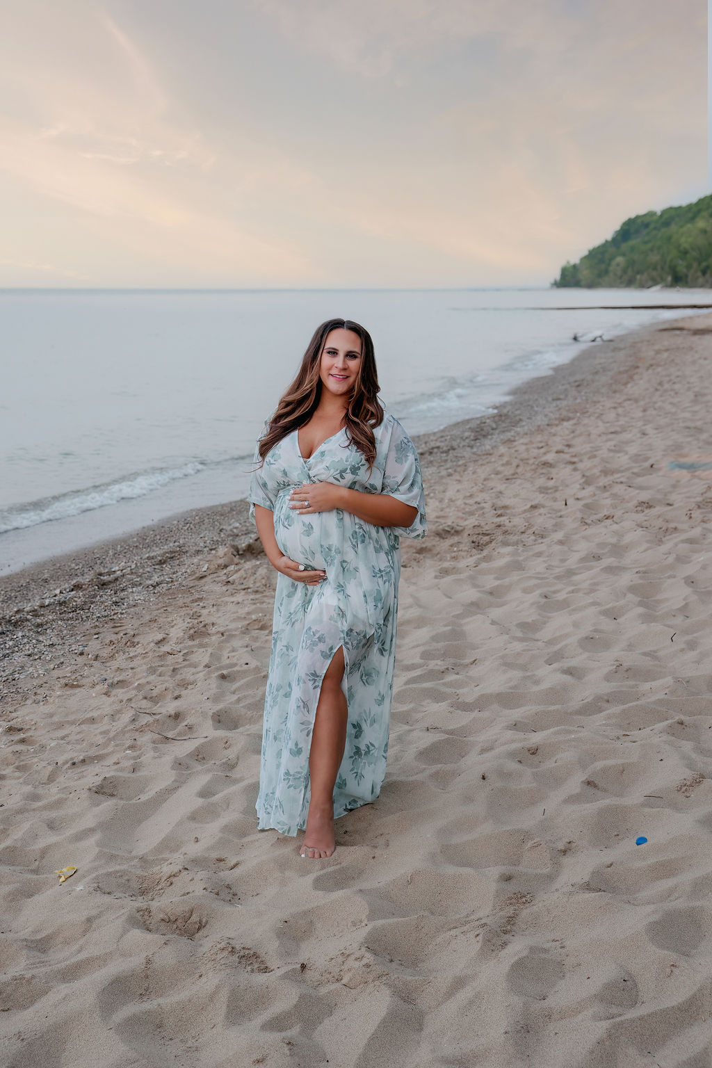 A mother to be in a blue floral dress walks on the beach while holding the bump at sunset after some birthing classes milwaukee
