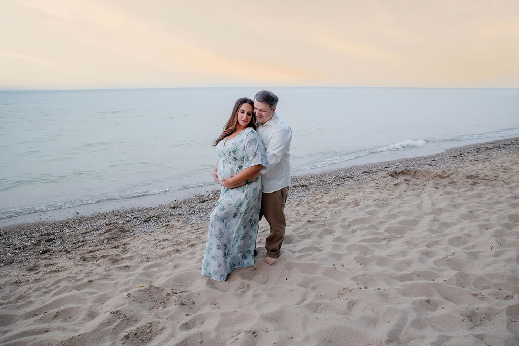 A mother to be leans into her husband while standing on a beach at sunset and holding the bump after some birthing classes milwaukee
