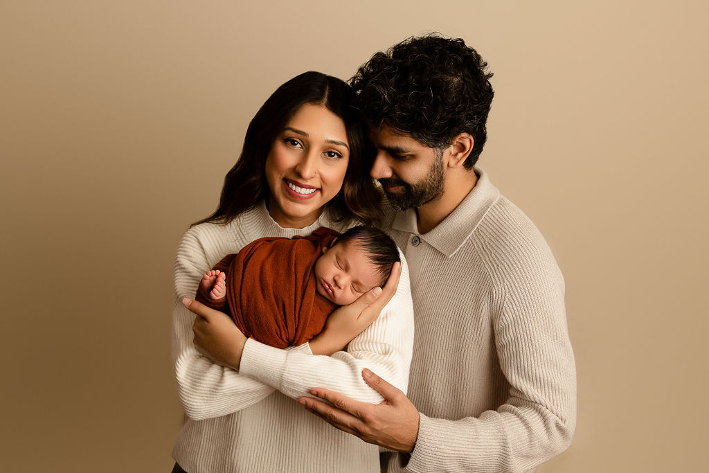 Happy parents cradle their sleeping newborn baby against mom's chest while standing in a studio