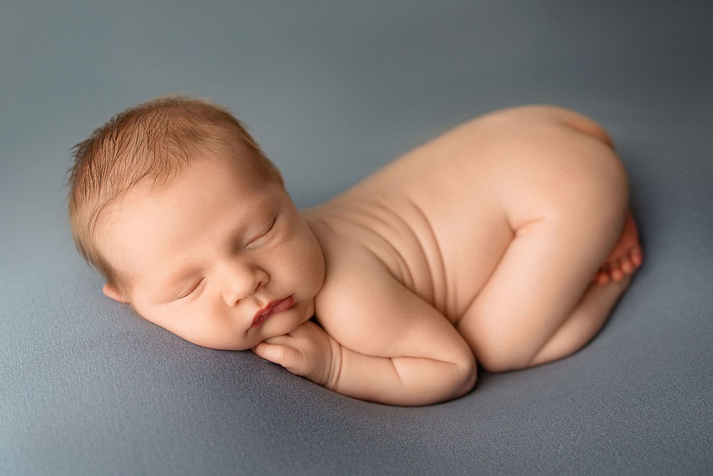 A newborn baby boy sleeps naked on a blue bed in a studio in froggy pose