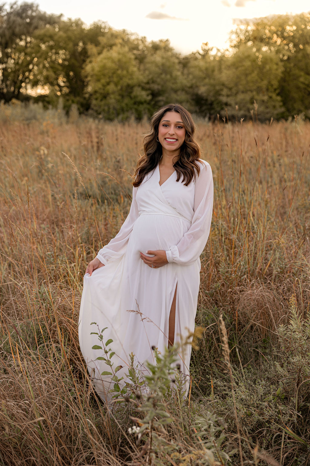 A mother to be holds her dress while walking through a field of golden grass at sunset
