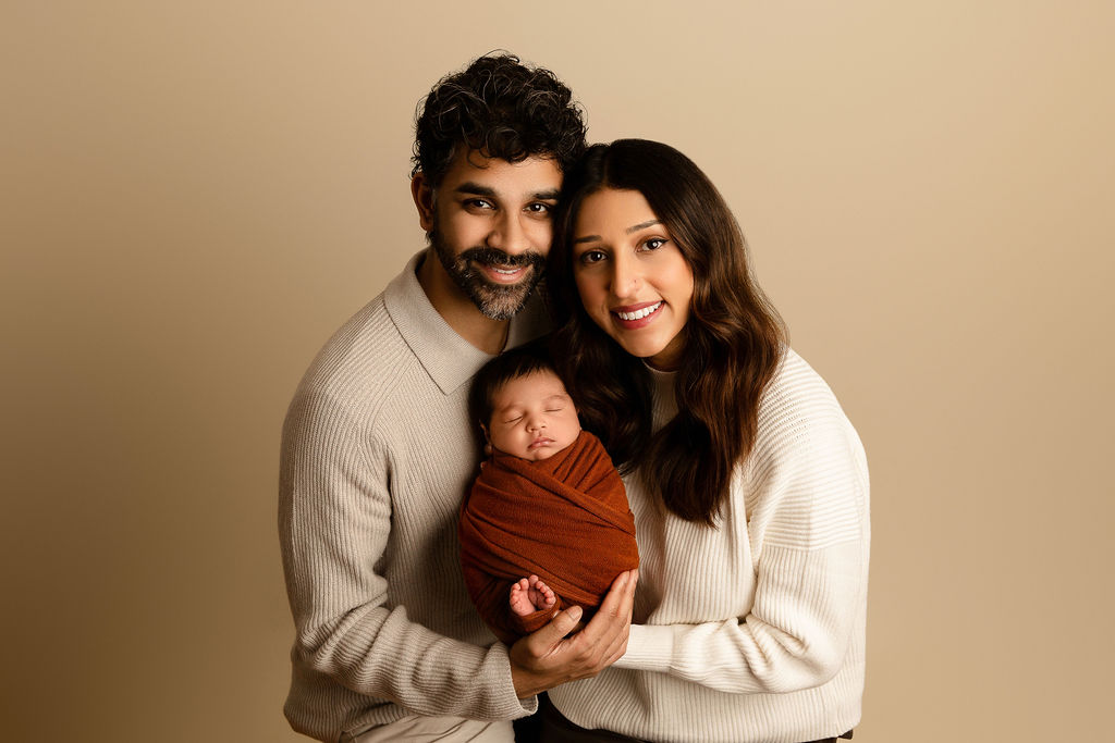 Happy parents smile while holding their sleeping newborn baby in a studio in a brown swaddle with toes sticking out