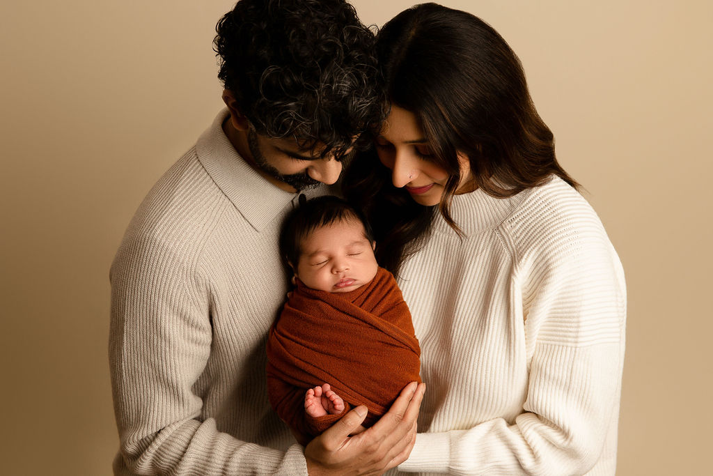 Happy parents in matching sweaters stand in a studio smiling down at their sleeping newborn baby in their hands between them after meeting float milwaukee