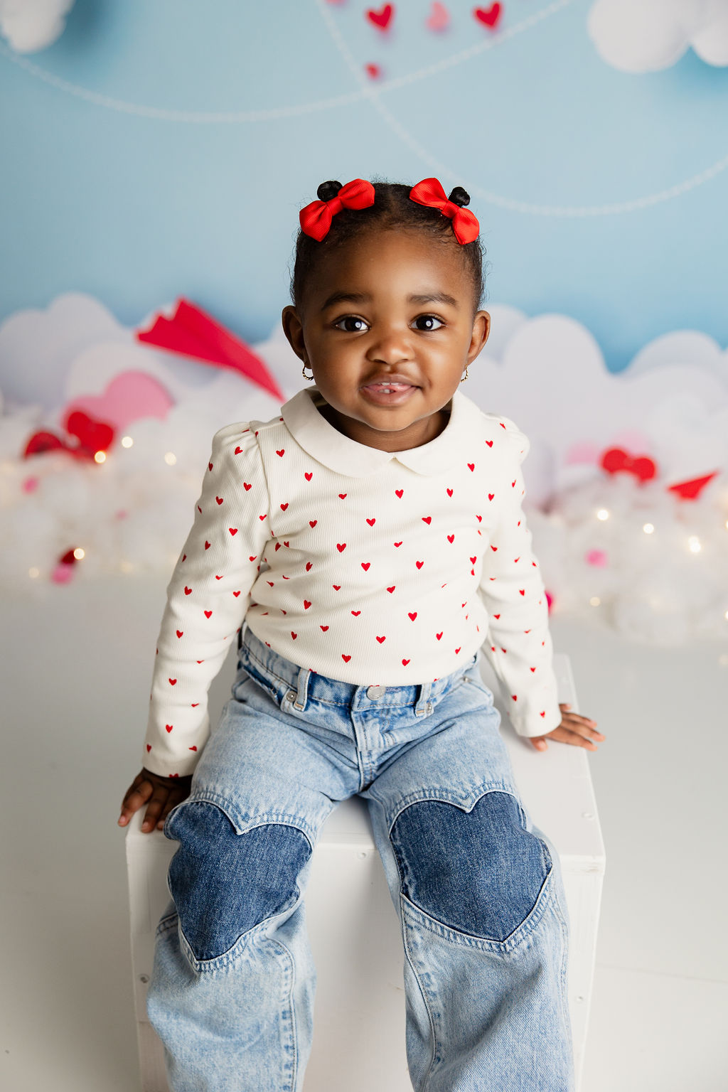 A toddler girl sits on a white box in a studio in jeans and a red heart polka dot shirt