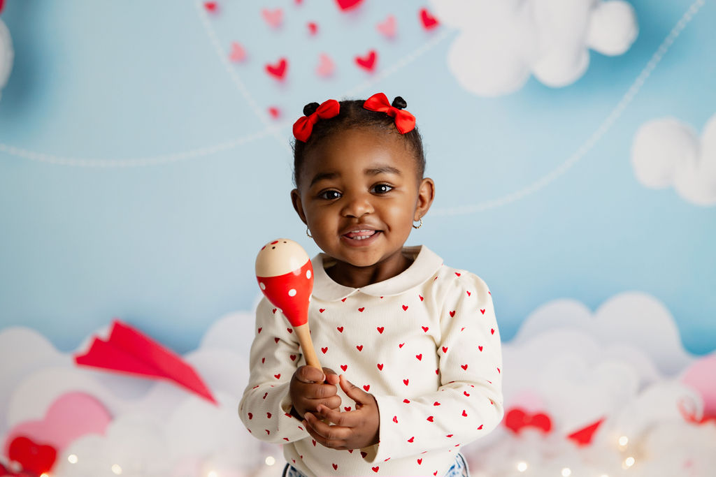 A happy toddler girl holds a red maraca in a white shirt with red hearts in a decorated studio after meeting florence eiseman milwaukee