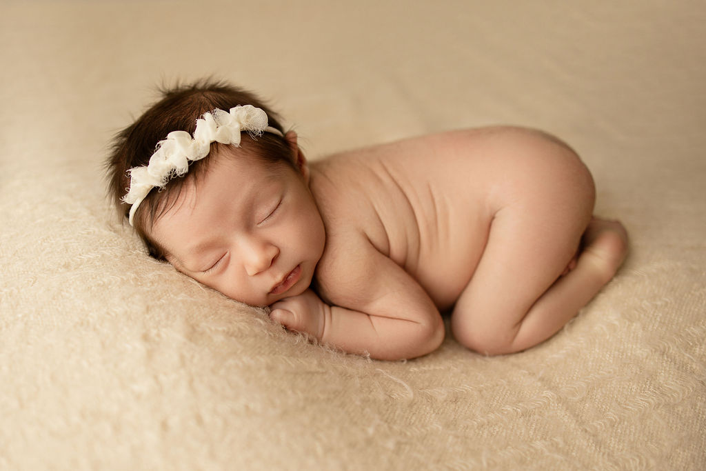 A newborn baby girl sleeps naked in a studio on a large pad with a headband