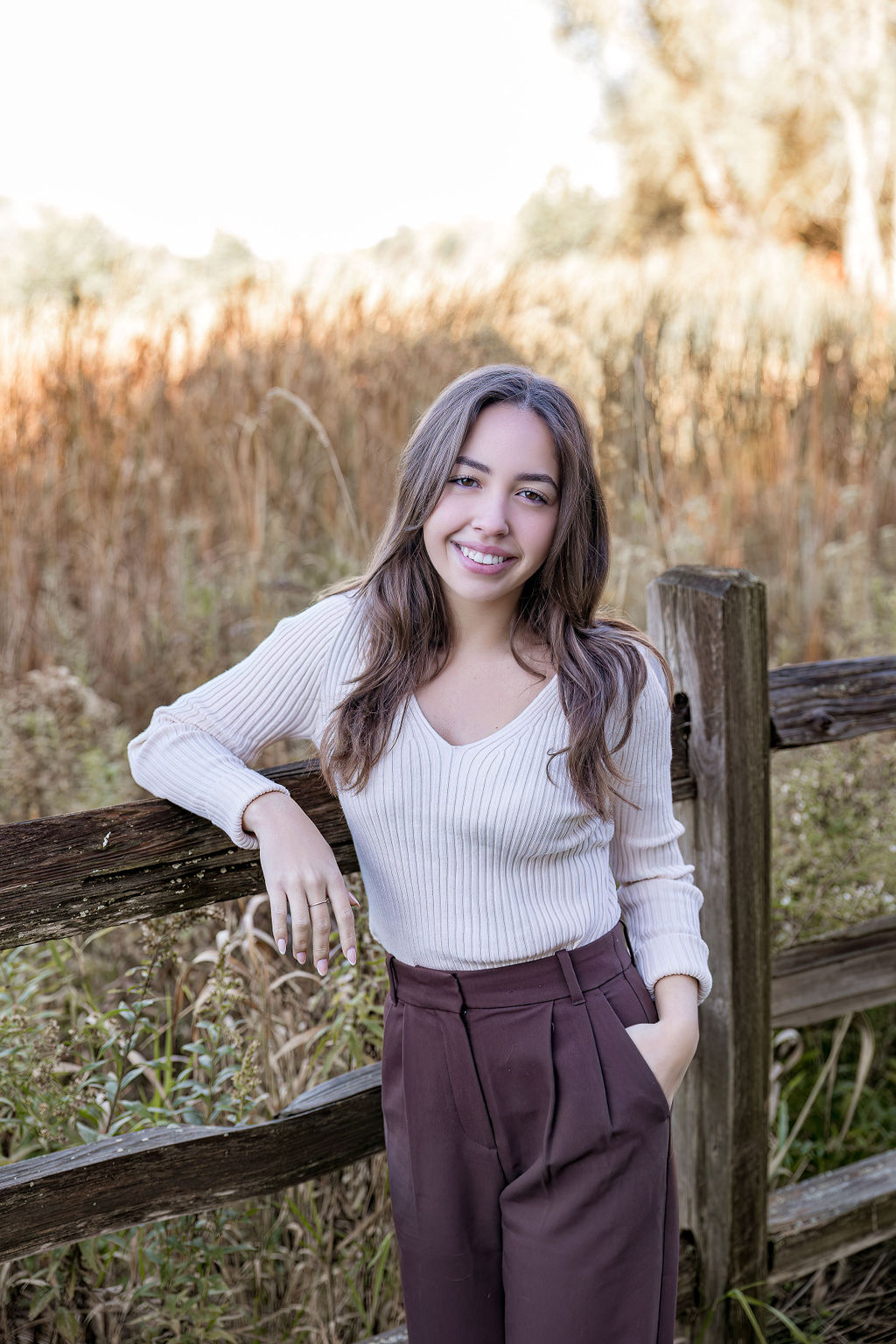 A teen girl in a white sweater leans against a wooden fence in a park