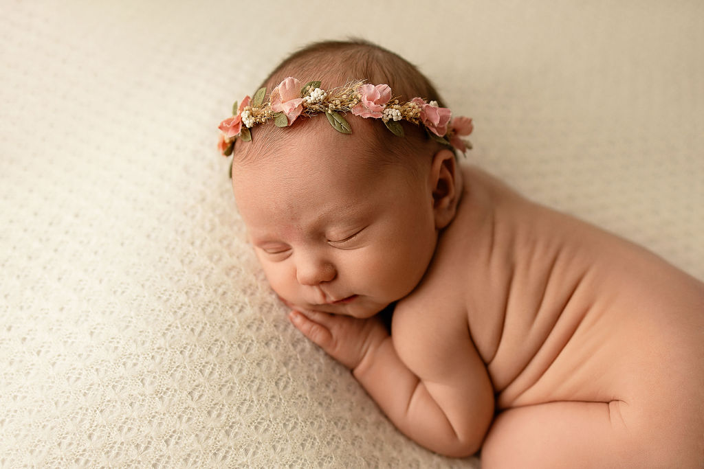 A newborn baby sleeps in froggy pose on a bed in a floral headband before some infant swim lessons milwaukee