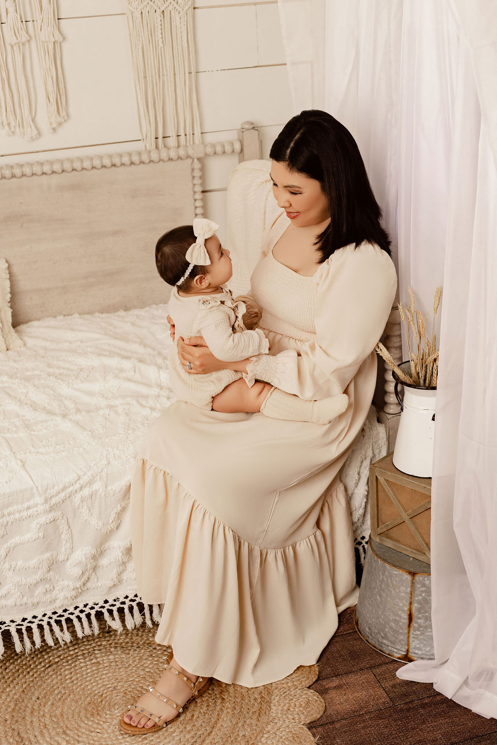 A happy mother in a cream dress sits on the edge of a bed in a studio smiling down at her infant daughter in her lap
