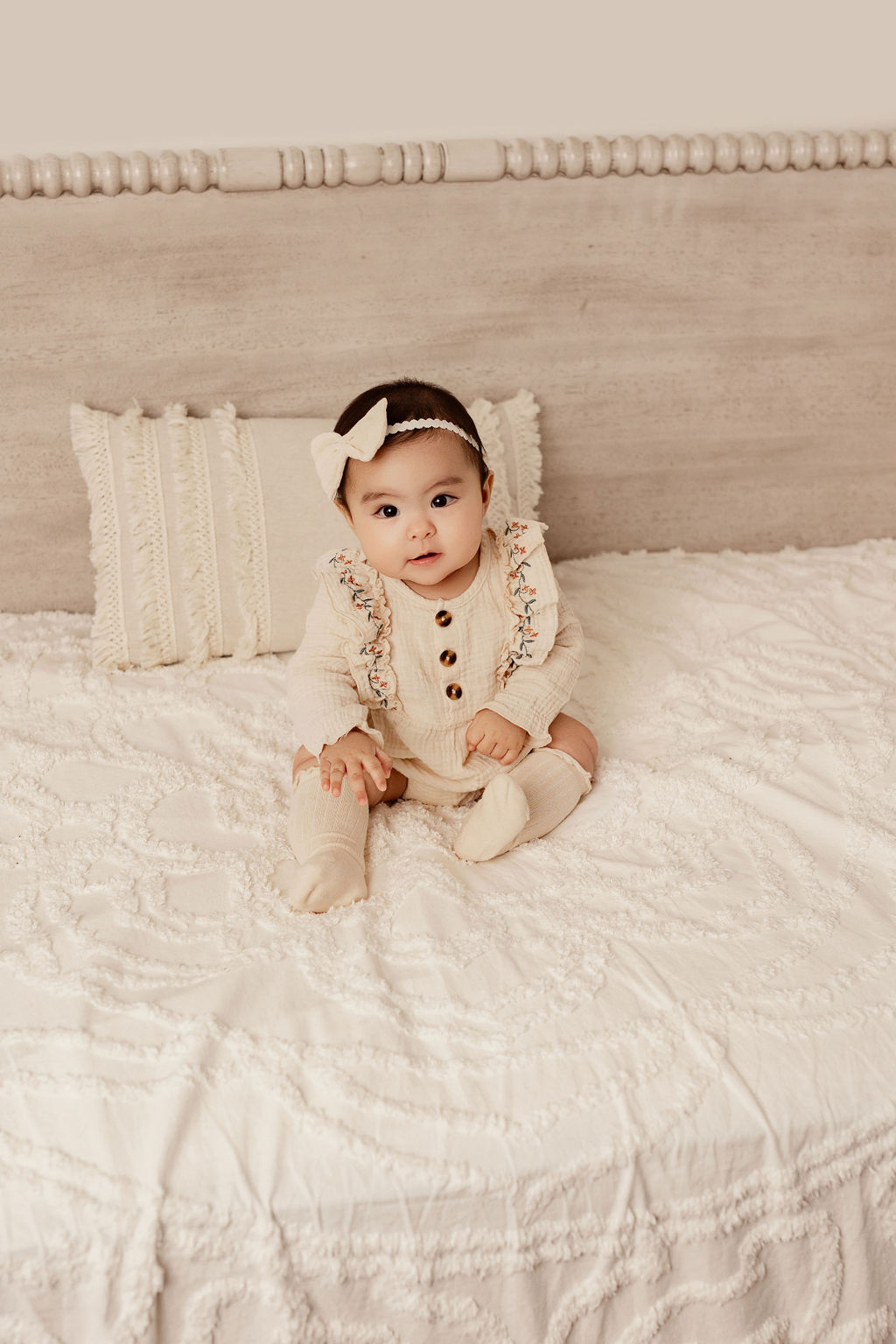 An infant girl in a cream onesie sits on a bed in a studio after meeting kenosha pediatricians