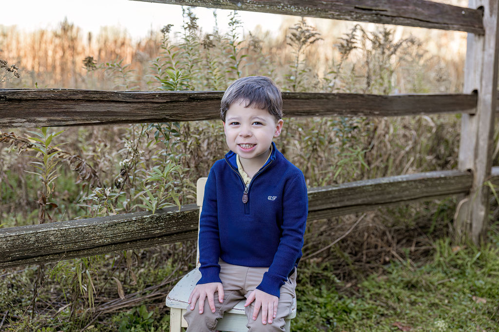 A happy toddler boy in a blue sweater sits on a wooden stool by a park fence