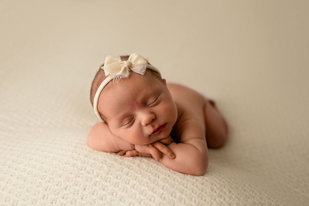 A newborn baby sleeps on her hands while laying on a white bed in a white bow