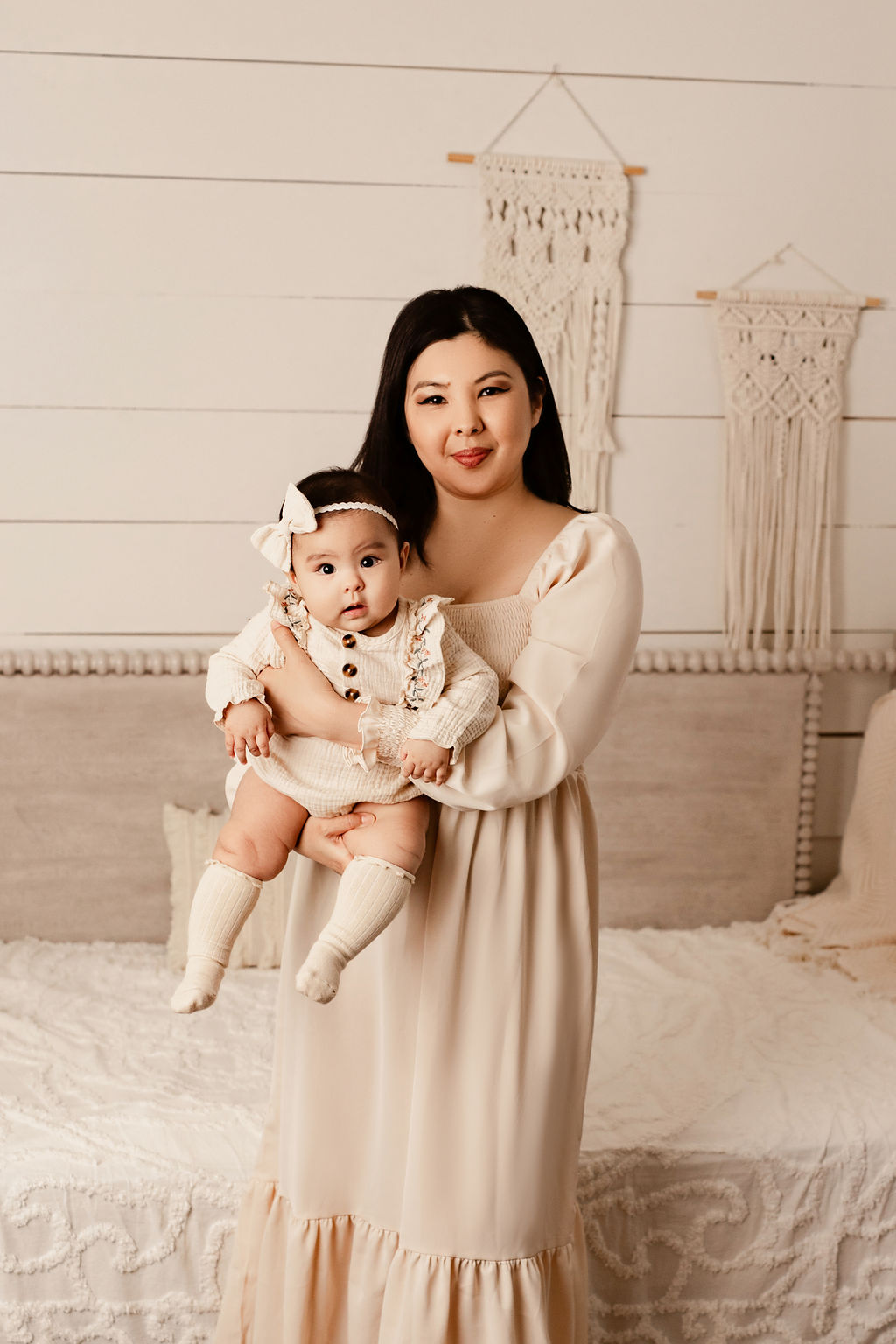 A happy mother in matching cream dresses stands in a studio bedroom holding her toddler daughter