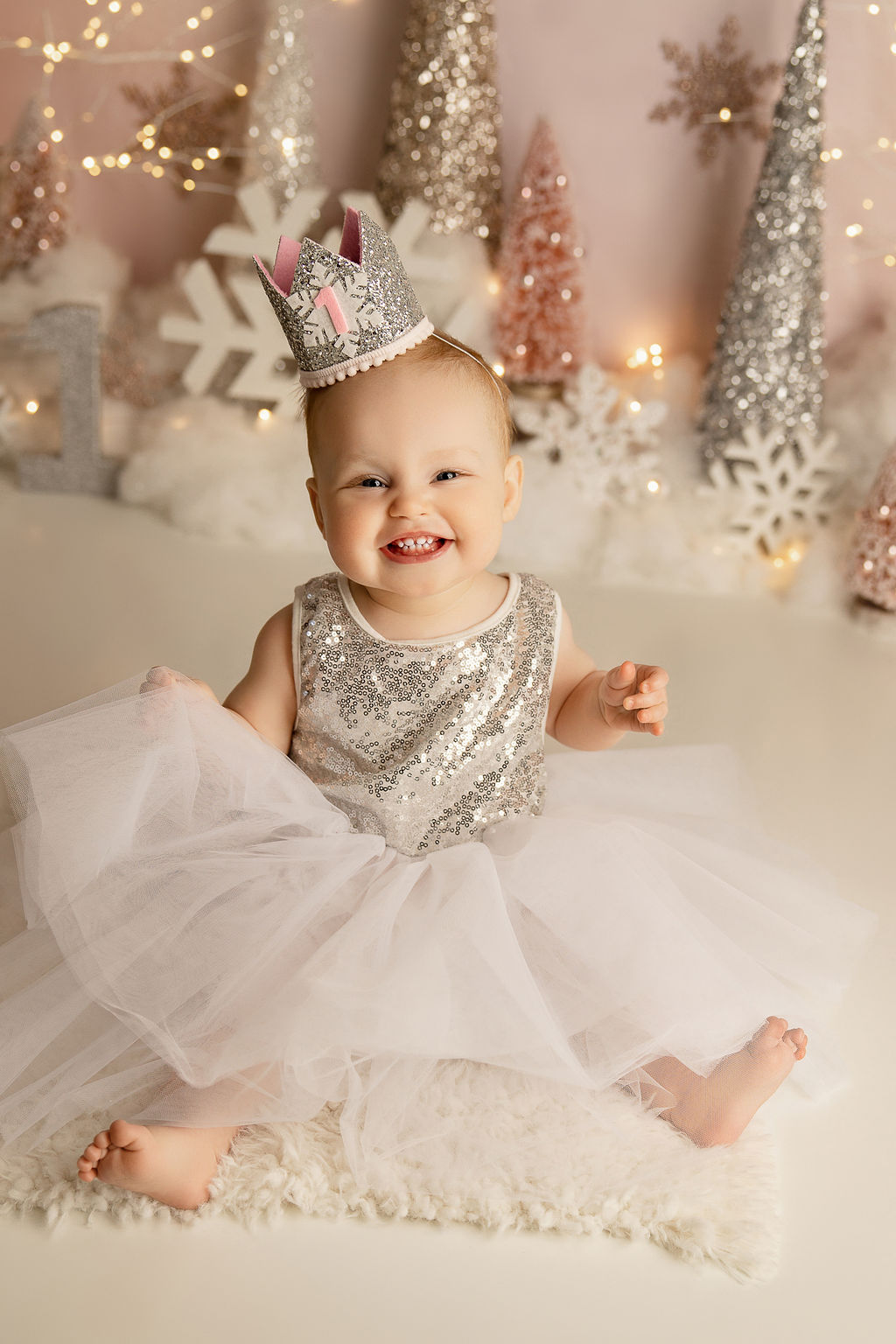 A toddler girl with a big smile sits on the floor of a studio in a silver sequence dress and crown