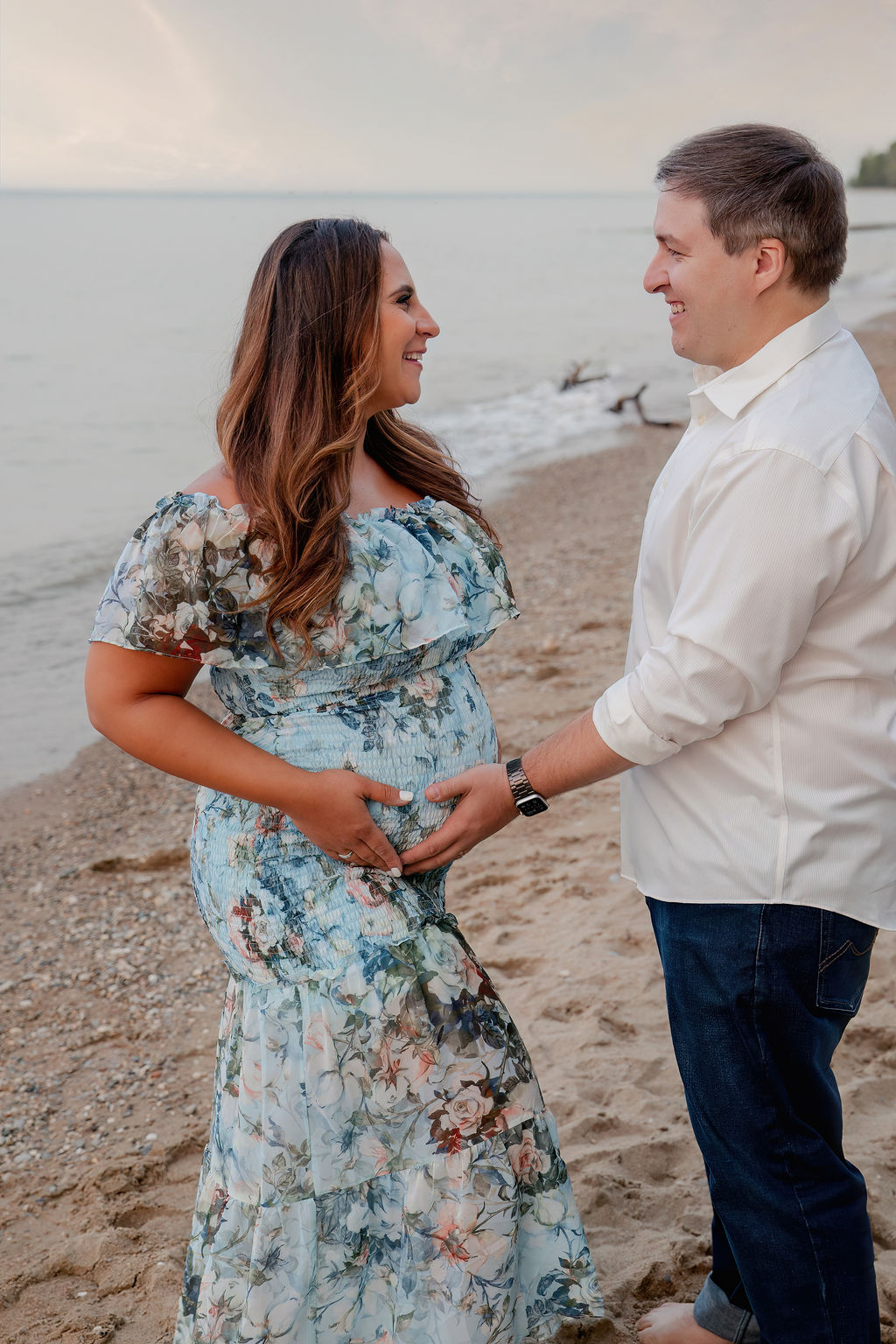 Expecting parents stand on a beach holding the bump and smiling at each other
