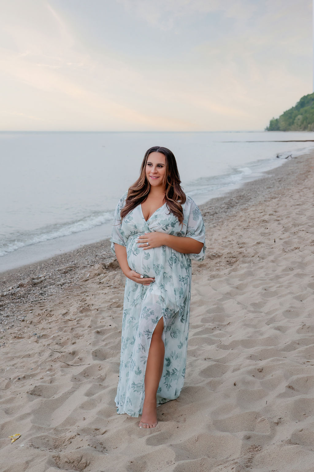 A mom to be walks down a beach at sunset in a blue floral print dress and holding her bump after visiting milwaukee baby shower venues