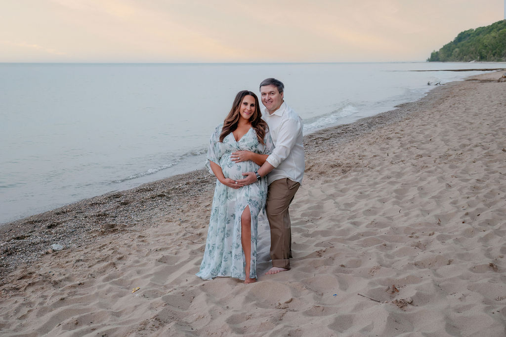 A mother to be in a blue floral dress stands on a beach holding her bump with her husband after visiting milwaukee baby shower venues