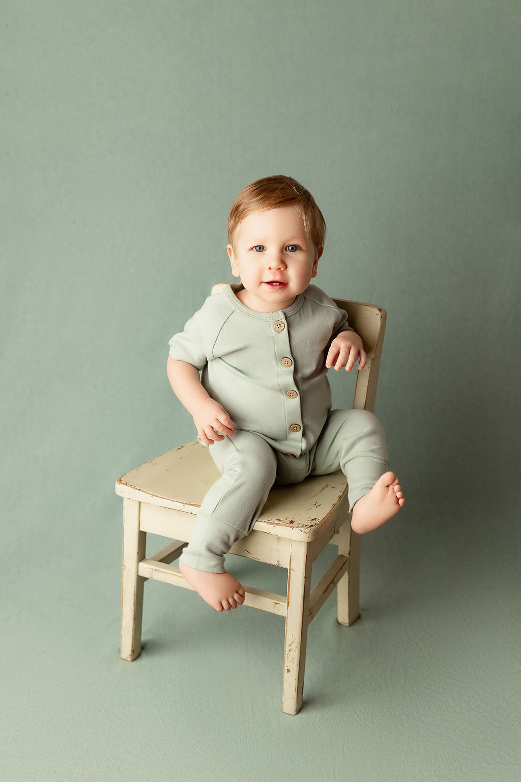 A toddler boy in a green onesie sits on a wooden chair