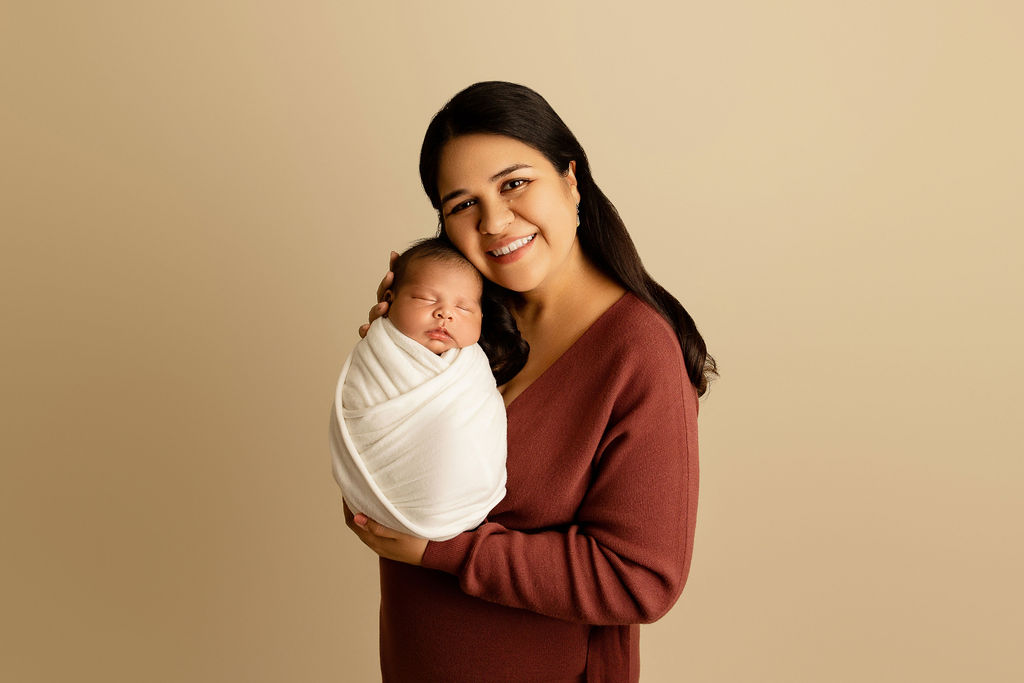 A happy mother in a pink sweater stands in a studio holding her sleeping newborn baby against her cheek