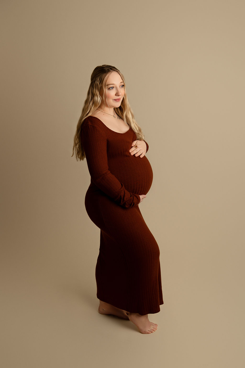 A mom to be holds her bump while standing in a studio in a red maternity gown