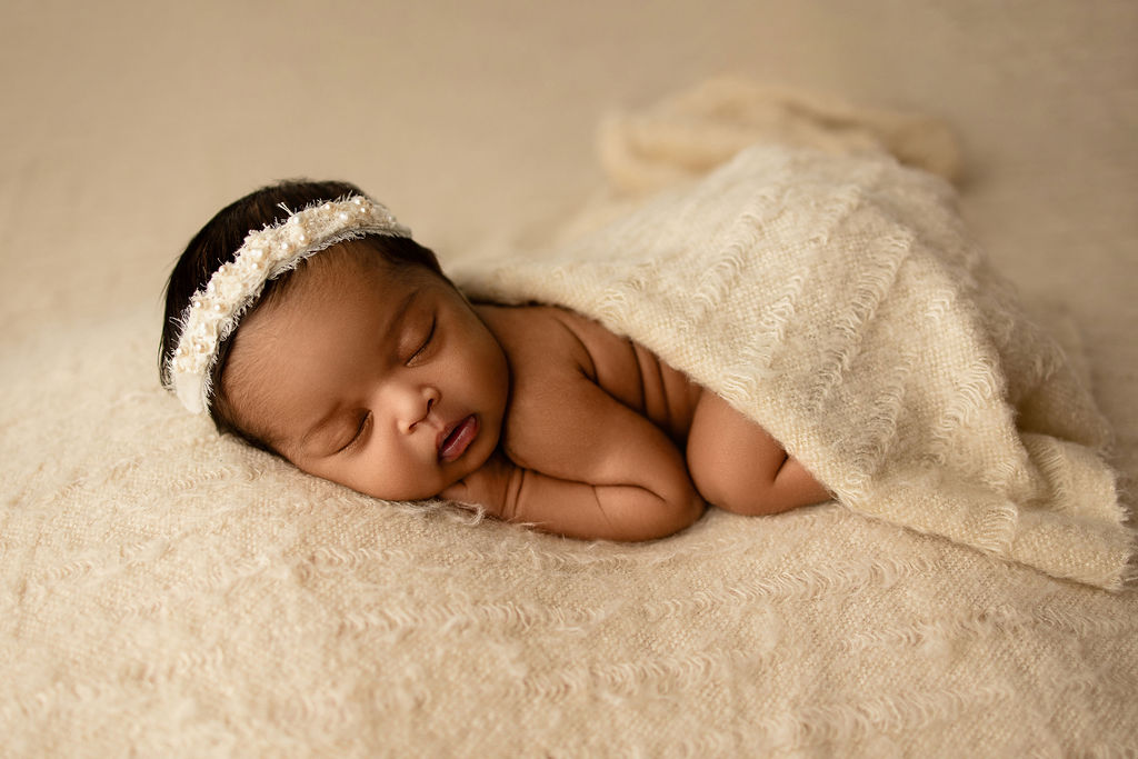 A newborn baby sleeps in froggy pose under a blanket with a white headband