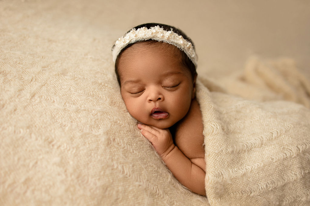 A newborn baby sleeps on her stomach under a blanket in a white headband thanks to milwaukee nannies