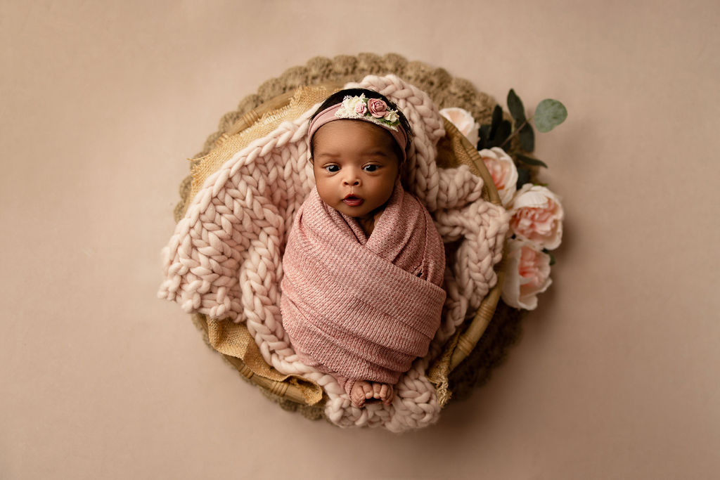 A newborn baby lays in a basket filled with blankets with eyes open in a pink swaddle after meeting milwaukee nannies