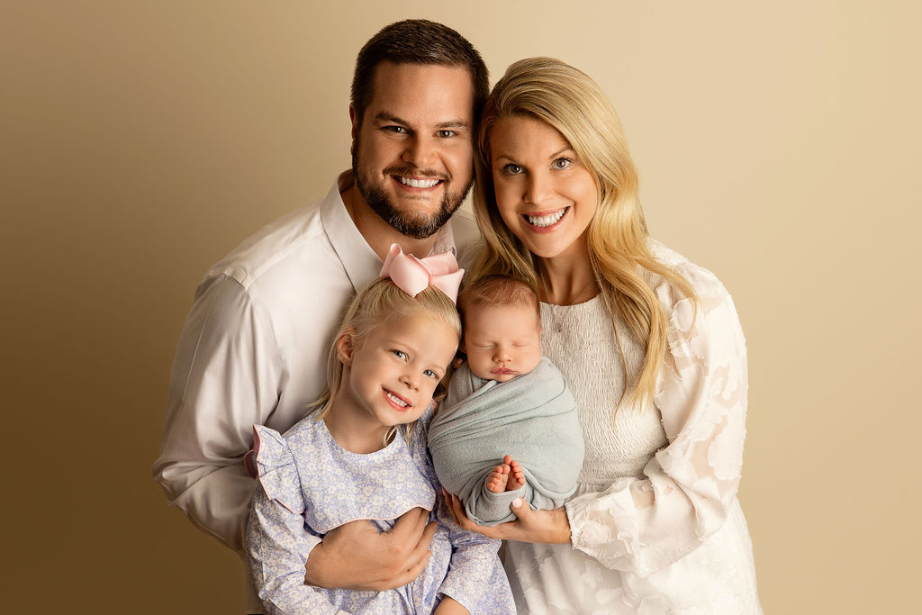 Happy parents smile while standing in a studio holding their sleeping newborn baby and toddler daughter thanks to milwaukee obgyn
