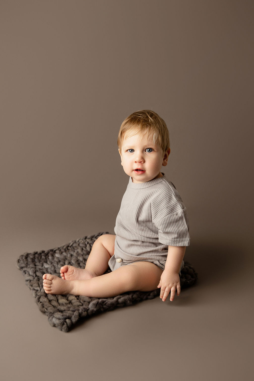 A young blonde toddler boy sits on a small mat in a studio in a grey onesie