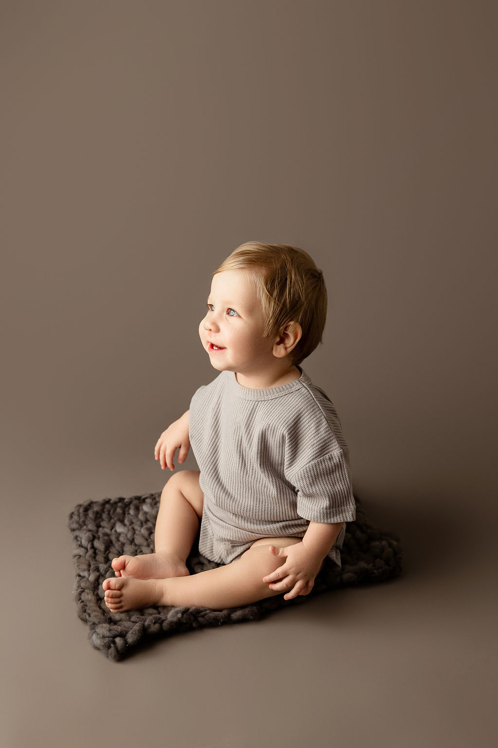 A toddler boy sits in a studio in a grey onesie smiling after meeting milwaukee pediatricians