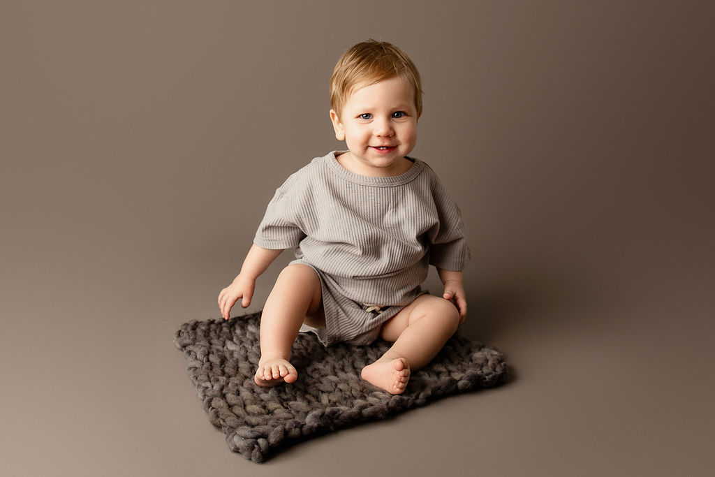 A young toddler sits on a small rug in a grey onesie in a studio after meeting milwaukee pediatricians