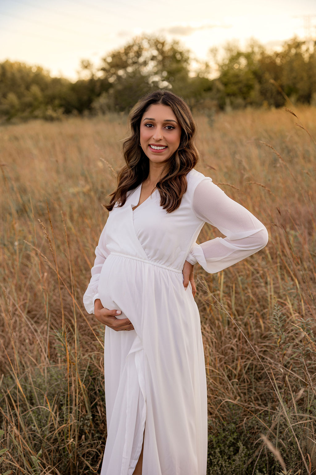 A mom to be in a white maternity gown holds her bump while smiling in a field of tall grass