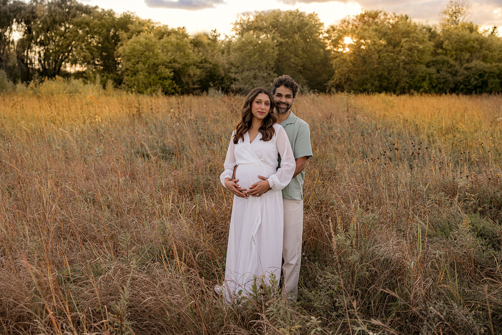 Happy expecting parents stand in a field of tall grasses at sunset holding the bump before some milwaukee prenatal yoga